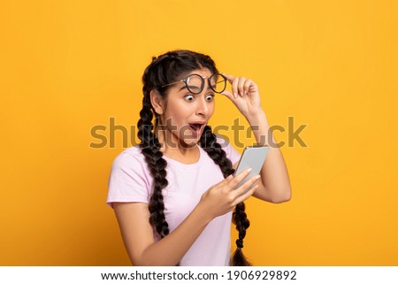 Wow, Great News. Portrait Of Excited Surprised Indian Woman Holding And Using Smartphone, Browsing Social Media. Emotional Young Lady Having Success, Taking Off Glasses, Yellow Studio Background Royalty-Free Stock Photo #1906929892