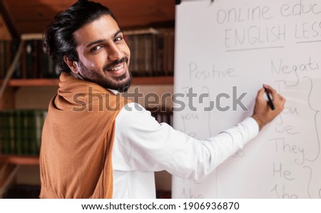 Cheerful Arab Teacher Guy Writing English Rules On Blackboard Having Online Lecture Indoor, Smiling To Camera. E-Learning And E-Teaching, Modern Virtual Education Concept