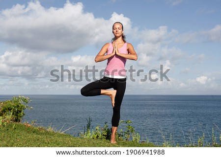 young attractive smiling blonde woman in sportswear doing yoga standing on the grass with blue sea and sky on the background