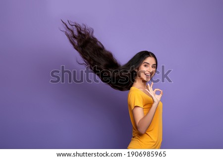 Hair Care Concept. Portrait of smiling indian model woman posing with long flying hair and showing okay sign gesture. Beautiful young lady standing isolated over purple studio background, copy space