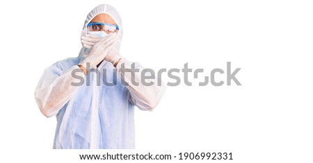 Young hispanic man wearing doctor protection coronavirus uniform and medical mask shocked covering mouth with hands for mistake. secret concept. 