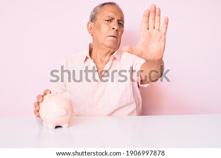 Senior handsome man with gray hair holding piggy bank sitting on the table with open hand doing stop sign with serious and confident expression, defense gesture 