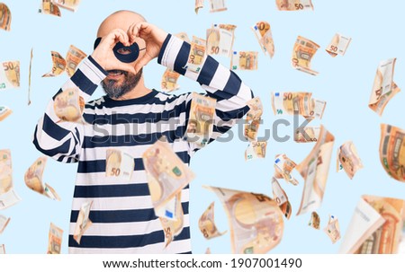 Young handsome man wearing burglar mask doing heart shape with hand and fingers smiling looking through sign
