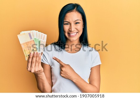 Beautiful hispanic woman holding south korean won banknotes smiling happy pointing with hand and finger 