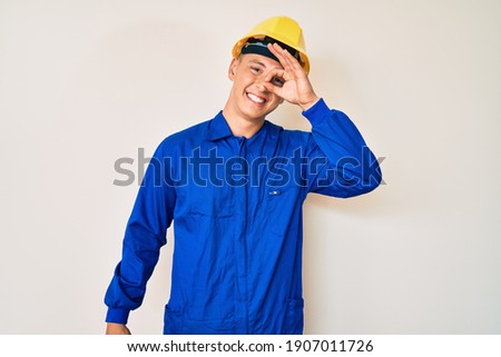 Young hispanic boy wearing worker uniform and hardhat smiling happy doing ok sign with hand on eye looking through fingers 