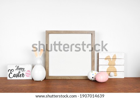 Mock up wood frame with Easter decor on a wood shelf. Shabby chic wood signs, eggs, modern glass bunny. Square frame against a white wall. Copy space.