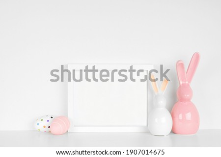 Mock up white frame with Easter Eggs and modern glass bunny decor on a shelf. White color scheme. Landscape frame orientation.