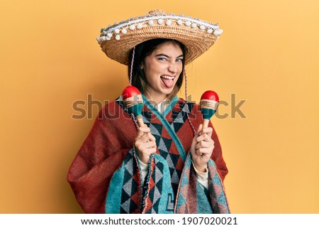 Young caucasian woman holding mexican hat using maracas sticking tongue out happy with funny expression.  Royalty-Free Stock Photo #1907020021