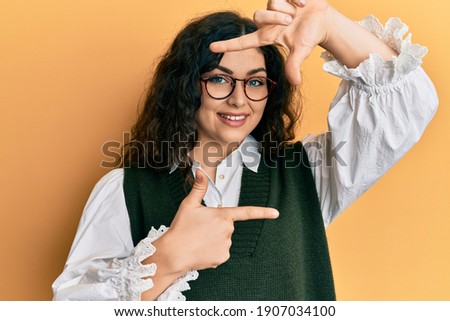 Young brunette woman with curly hair wearing casual clothes and glasses smiling making frame with hands and fingers with happy face. creativity and photography concept. 