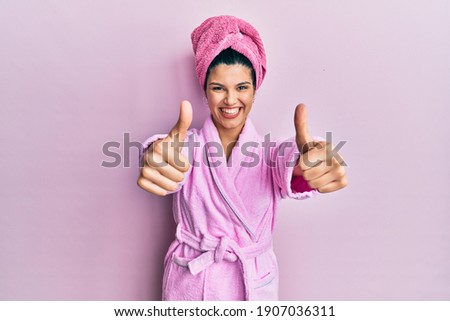 Young hispanic woman wearing shower towel cap and bathrobe approving doing positive gesture with hand, thumbs up smiling and happy for success. winner gesture. 