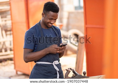 young african carpenter smiling while using his phone Royalty-Free Stock Photo #1907037448