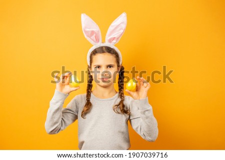 Happy Easter, kids. A girl in rabbit ears on the head with painted eggs on a yellow background. Funny smiling child