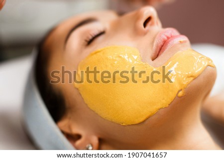 Procedure for skin care. Doctor in cosmetology office applies an yellow gold alginate mask to the face of young pretty woman. Royalty-Free Stock Photo #1907041657