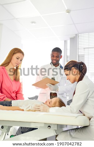 team of doctors working together while treating kid girl patient lying on hospital bed, african man and caucasian woman consulting girl and holding medical check-up