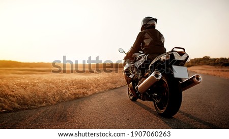 Handsome motorcyclist in leather jacket and helmet at sunset on the road in warm sun rays Royalty-Free Stock Photo #1907060263