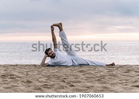 Modern healthy Man with beard in white clothes doing stretching yoga exercises in the beach
