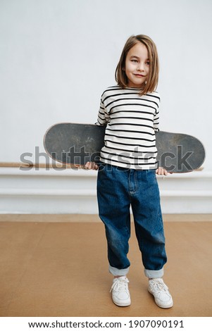Thougthful little girl posing with skateboard for a photo with a tired sad fake smile. Holding it behind her back. She wears striped black and white shirt. At indoor skatepark in winter time.