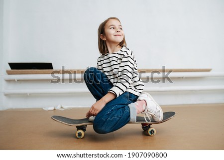 Pretty little girl in striped black and white shirt sitting on one knee on a skateboard with style, posing for a photo, looking away. At indoor skatepark in winter time.