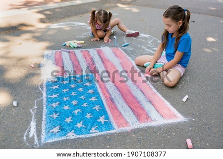 Two cute friends girls  drawing American flag with colored chalks on the sidewalk near the house on sunny summer day. Kids painting outside. Creative development of children.