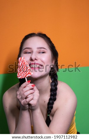 charming brunette woman in yellow top holding red big lollipop on bright green and orange background. sweets concept. candy. copy space