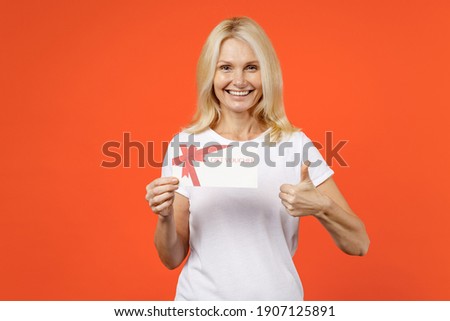 Cheerful elderly gray-haired blonde woman lady 40s 50s years old wearing white basic t-shirt hold gift certificate showing thumb up looking camera isolated on orange color background studio portrait