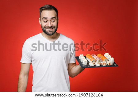 Excited young bearded man 20s wearing casual white t-shirt biting lips hold in hand makizushi sushi roll served on black plate traditional japanese food isolated on red background studio portrait