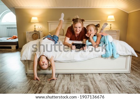 Mother and her daughters spending time at home watching movies, cartoons or serials on the tablet. Girls are laying on the bed, one girl got bored and having fun by herself. Family weekend. 