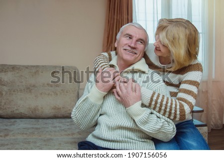 Happy positive senior elderly couple hugging while sitting on the sofa. Middle-aged people talk, laugh, relax, have fun together at home. Place for text. Royalty-Free Stock Photo #1907156056