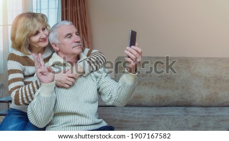A modern elderly couple, 50-60 years old, is sitting on the couch at home, taking a selfie on a smartphone in the living room. Husband and wife are having fun by video calls. Copyspace