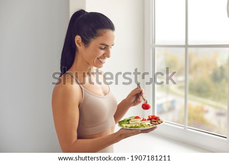 Smiling pretty young fitness woman standing and eating balanced healthy meal before or after workout at home window. Healthy diet, active fitness lifestyle, clean eating food concept