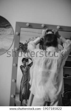 Black and white photo of girl in long dress making photo of lady in transparent blouse in mirror in apartment. Two women taking pictures inside. Blurred effect.