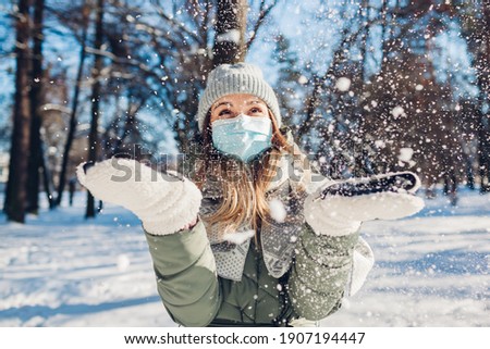 Winter portrait of happy woman in protective mask playing with snow outdoors. Corona virus covid-19 pandemic new reality. Girl having fun throwing snow