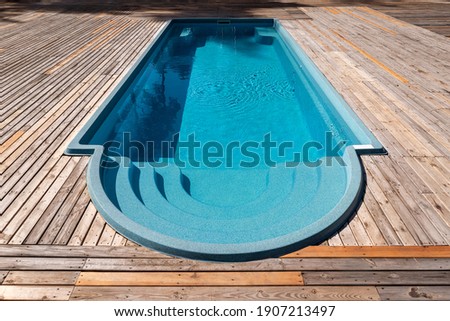 New modern fiberglass plastic swimming pool entrance step with clean fresh refreshing blue water on bright hot summer day at yard or resort hotel spa area. Wooden flooring deck of teak or larch board Royalty-Free Stock Photo #1907213497