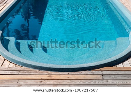 New modern fiberglass plastic swimming pool entrance step with clean fresh refreshing blue water on bright hot summer day at yard or resort hotel spa area. Wooden flooring deck of teak or larch board Royalty-Free Stock Photo #1907217589