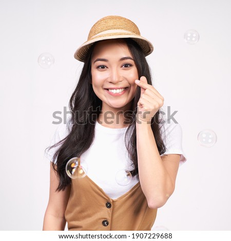 Portrait of a charming lovely lady in a white T-shirt and straw hat, makes a Korean heart sign, expresses her love and sympathy, posing on a white background. Body language concept.