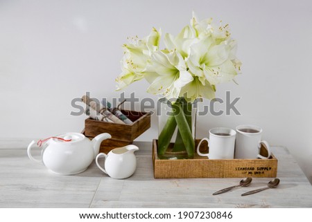 A bouquet of white lily in a glass vase on a table with two tall cups of coffee, a teapot, spoons, and a milk jug. Copy space