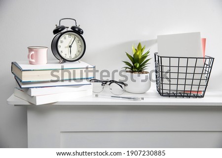 A stack of books, an artificial succulent plant in a ceramic pot, glasses, a metal basket with notebooks, a pen and a cup on a white table surface. Concept - office at home. Online training.