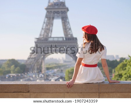 A beautiful girl in a white dress, a red beret sits against the backdrop of the Eiffel Tower in Paris. Travel to Paris. Ideas for a photo shoot. Royalty-Free Stock Photo #1907243359