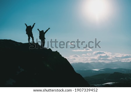 Silhouettes of two happy hikers in winner poses with raised arms are standing on mountain top