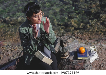 Young Indian girl reading a book near lake with a lamp and orange besides her , wearing green jacket