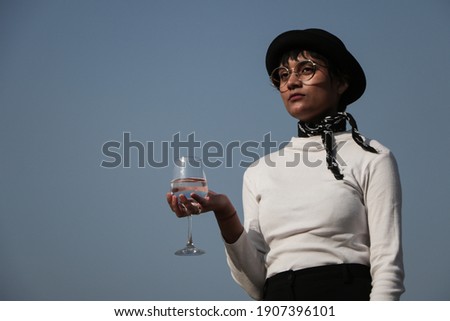 A portrait of an young indian girl holding a wine glass in hand looking to the camera, wearing a hat and white top , blue sky behind subject