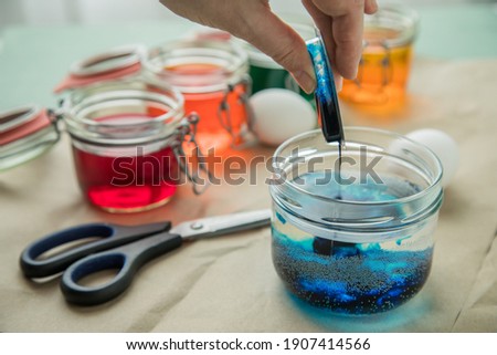 Woman pouring liquid orange Easter cold colour color in glass with water and vinegar next to green, yellow, red, blue dye in packaging with boiled eggs and scissors for DIY coloring