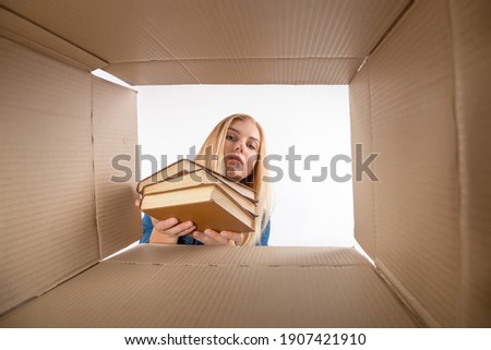 Young beautiful girl blonde, hold books, opens and peeks into cardboard box. To get or to put things to move, bottom view.