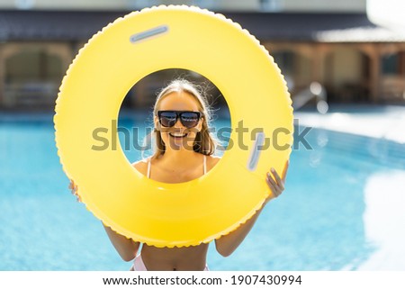 Beautiful woman in bikini holding a inflatable ring at poolside and smiling. Female wearing bikini and sunglasses enjoying her holiday at resort poolside.