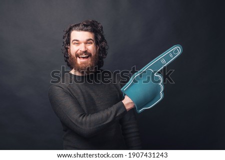 Picture of a man with beard is holding a fan glove pointing at a free space being excited is looking at the camera .