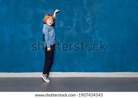 Wow look, empty place on wall for advertising. Amazed kid boy on tiptoe pointing to blank space on background. Surprised preschooler showing height on backdrop with copyspace for ad.