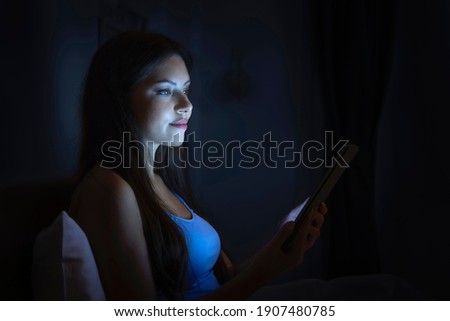 A beautiful woman in bed reads on her tablet device before going to sleep in a dark bedroom