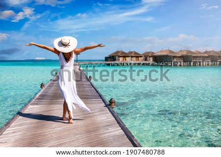 A happy woman in white summer dress and with raised arms walks over a wooden pier in the Maldives islands and enjoys her tropical holidays Royalty-Free Stock Photo #1907480788