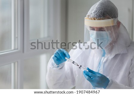 A doctor or healthcare professional in a personal protective kit, wearing rubber gloves in a hospital room in an epidemic or pandemic of coronavirus or COVID 19, draws a syringe with medicine. Vaccine