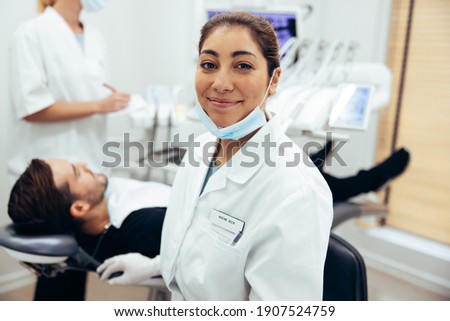 Happy female dentist looking at camera. Dental doctor in her clinic with patient in dentist's chair and nurse in background. Royalty-Free Stock Photo #1907524759